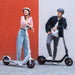 Two people standing with electric scooters against a red and gray wall.