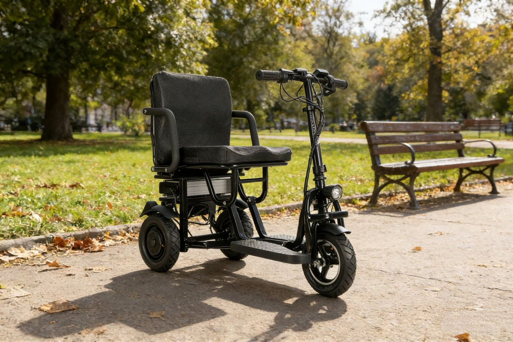 Black electric wheelchair on a paved path in a park with trees and a bench in the background.