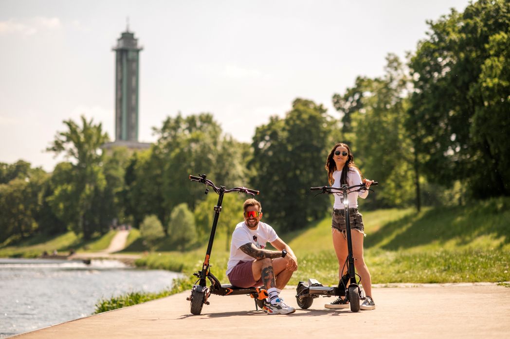 Two people with electric scooters in a park setting with trees and a tower in the background.