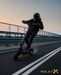 A person wearing a black helmet and black outfit rides a black electric scooter with gold accents on a road bridge during sunset. The brand name 'ROLEY X' is visible in the bottom right corner.