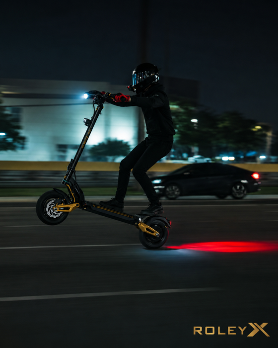 A person in black riding apparel and a helmet maneuvers a black electric scooter on a dark nighttime road, with the scooter's red taillight glowing on the pavement. The word ROLEXY appears in the bottom right corner.