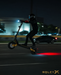 A person in black riding apparel and a helmet maneuvers a black electric scooter on a dark nighttime road, with the scooter's red taillight glowing on the pavement. The word ROLEXY appears in the bottom right corner.